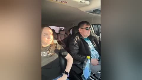 Video of a woman with dark hair, wearing a black shirt, and a man in sunglasses and a black jacket, both seated in a car, with a blurred background.