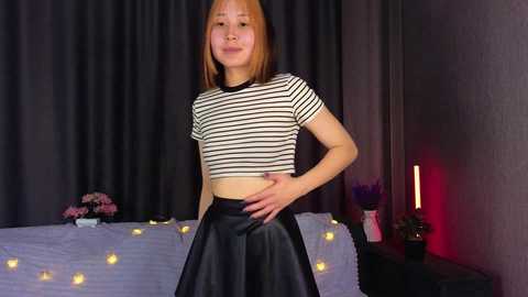 A video of an Asian woman with straight, shoulder-length auburn hair, wearing a striped crop top and black leather skirt, standing in a dimly lit bedroom with dark curtains, fairy lights, and a lit candle.
