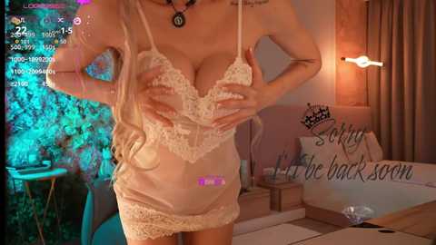 A video of a slender, blonde woman in a lacy, sheer, white lingerie dress with a purple vibrator in her hand, standing in a dimly lit, cozy bedroom.