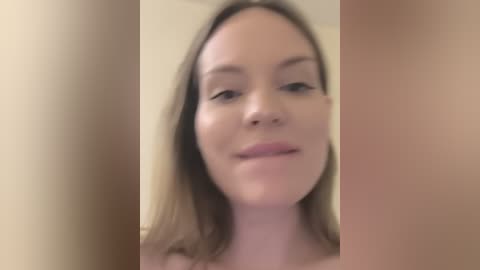 A blurred video of a fair-skinned woman with straight, shoulder-length blonde hair and a neutral expression, set against a beige background.