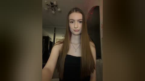 Video of a young Caucasian woman with long straight brown hair, fair skin, and light makeup. She wears a black strapless top and a thin necklace. Background shows a dimly lit room with a wardrobe, chandelier, and door frame.