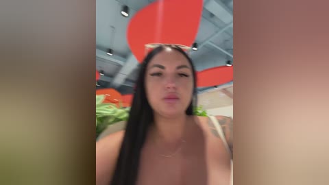 A video of a woman with long black hair and light skin, wearing a white top, blowing a kiss. Background features red and orange circular decorations and green plants.