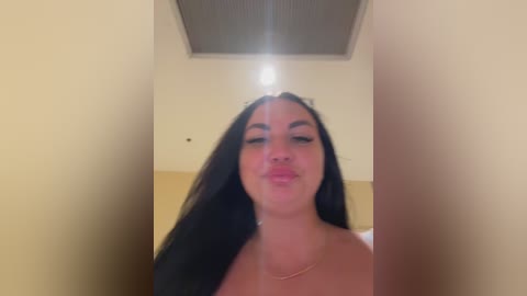 Video of a young woman with long, straight black hair, medium skin tone, and full lips, wearing a strapless top, standing indoors with a beige wall and ceiling light fixture in the background.