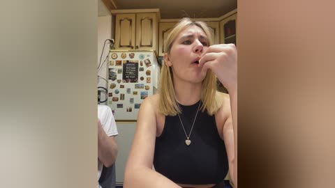 Video of a blonde woman in a black sleeveless top, with a pendant necklace, eating a cookie in a kitchen with wooden cabinets and a collage of photos on the wall.