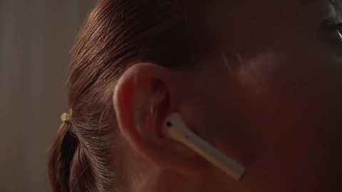 Close-up video of a person with light brown hair, wearing a white earbud, with a blurred, warm-toned background. The image focuses on the ear and neck, highlighting the soft, smooth texture of the skin.