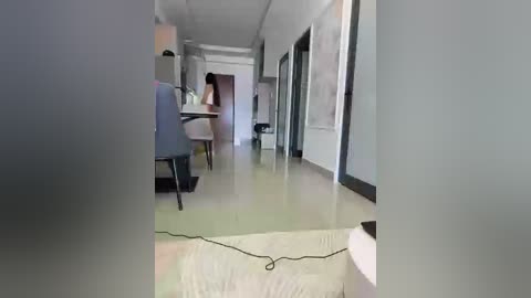 A video of a modern, minimalist hallway with light-colored walls, a beige floor, and gray chairs. A person wearing a white shirt and dark pants walks towards the camera, partially obscured.