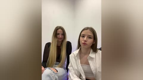 Video of two young women with long blonde hair, one in a black top, the other in a white shirt. Both have light skin and appear casual.