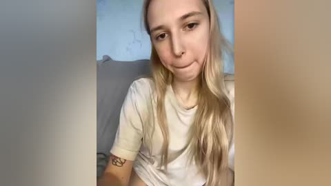 A close-up video of a young woman with long blonde hair, pale skin, and a small tattoo on her forearm, wearing a white t-shirt, smiling slightly, sitting on a couch with light-colored walls in the background.