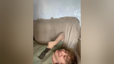 Video of a young man with light skin, brown hair, and a green shirt, laughing, lying on a beige couch, surrounded by white walls with floral patterns.