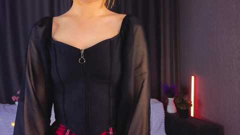 Video of a woman in a black sheer blouse, showing off her cleavage, with a red plaid skirt, standing in a dimly lit bedroom with a red lamp and floral decor.