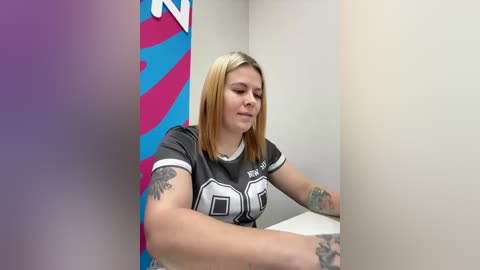 Video of a young woman with blonde hair, wearing a gray football jersey, sitting in a bathroom with colorful, abstract wall art and a tattoo on her arm.