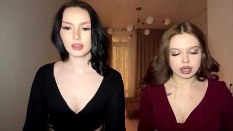Video of two young women with fair skin, one with black wavy hair and a black V-neck top, the other with brown wavy hair and a burgundy top. They stand in a softly lit room with beige walls and large windows.