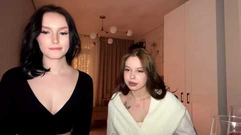 Video of a tall, slender woman with long black hair in a black V-neck top standing beside a petite, brunette woman in a white robe.