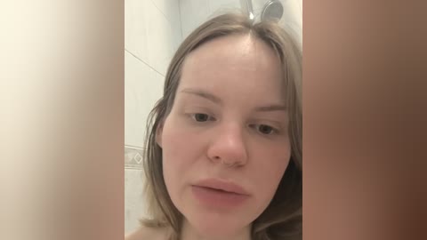 A video of a fair-skinned woman with shoulder-length brown hair, wearing a nose ring, and looking directly at the camera in a tiled bathroom, with her face framed by two vertical, beige bathroom walls.