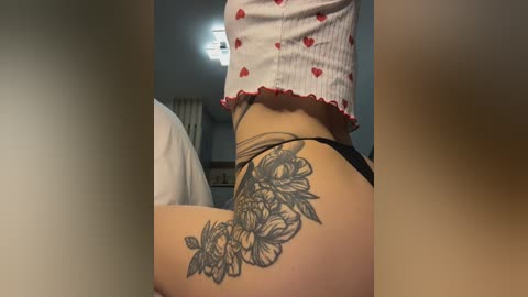 Video of a person with light skin, wearing a white crop top with red hearts, and black thong, displaying a large, intricate floral tattoo on the right hip. Background shows a dimly lit room with a bed and curtains.