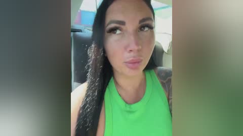 A video of a young woman with long, straight black hair and a tan complexion, wearing a neon green sleeveless top. She has a serious expression, and her makeup is subtle. The background shows a car interior.