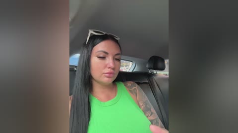 A video of a tattooed woman with long black hair wearing a neon green top, sitting in a car with sunglasses on her head, looking down.