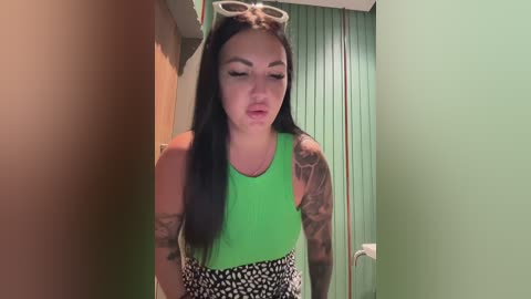 Video of a young woman with long black hair, wearing a neon green tank top and black and white skirt, standing in a hallway with green walls.