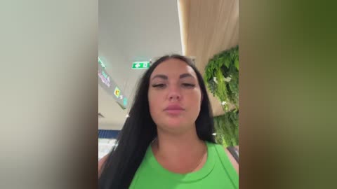A video of a woman with long black hair, wearing a bright green tank top, standing in a shopping mall with green plants in the background.