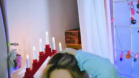 Video of a woman with brown hair in a light blue shirt, resting her head on a white pillow. Red candle holders with lit candles are on a table, surrounded by white curtains.
