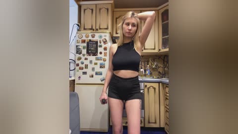 Video of a slim, blonde Caucasian woman in a black sports bra and shorts, standing in a cluttered kitchen with beige cabinets, a fridge covered in photos, and a sink.