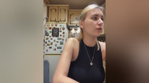 A video of a blonde woman in a black sleeveless top, wearing a heart pendant necklace, standing in a cozy, wooden-paneled kitchen adorned with colorful magnets and a framed quote on the fridge.