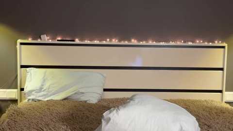 Video of a modern bedroom featuring a white wooden headboard with a black horizontal strip, lit by warm, white fairy lights. A beige shag rug covers the bed, with white pillows and a white duvet. The walls are painted dark gray.