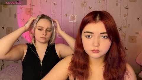 Video of two young women with fair skin in a bedroom. One with short blonde hair styles the other with long red hair. Background features butterfly-patterned wallpaper and a pink light.