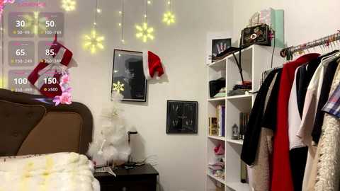 Video of a cozy bedroom with a white bed, a black nightstand, a Christmas tree, a white and red wreath, a framed picture, and a white shelf with clothing.
