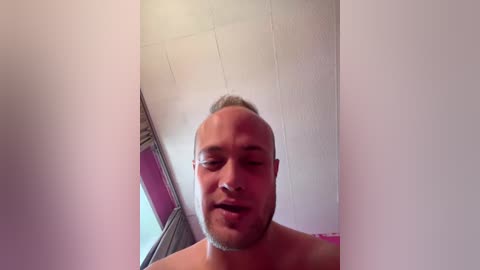 Video of a topless, bald, bearded man with light skin, slightly smiling, in a dimly lit room with a slanted ceiling.