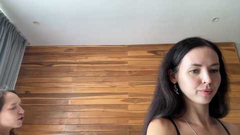 Video of a young woman with long black hair and fair skin, wearing a black top, standing in a room with wooden paneling and a gray curtain.