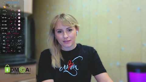 Video of a blonde woman with straight hair wearing headphones, a black t-shirt with \"Avon\" in red and white text, and a blurred background featuring a TV screen with financial data.