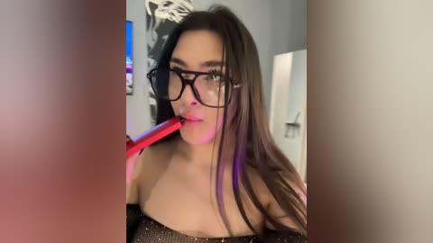 Video of a young woman with long dark hair, glasses, and a black glitter top, holding a red vibrator to her lips, in a dimly lit room with a TV and gray walls.