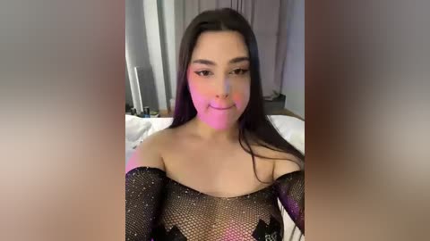 Video of a young woman with long dark hair, fair skin, and light makeup, wearing a black mesh top, sitting on a bed with white sheets, in a dimly lit bedroom.
