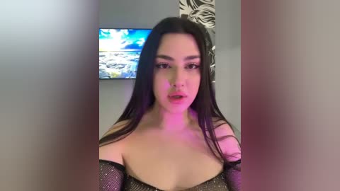 Video of a young woman with long, straight black hair, wearing a black, off-shoulder glittery dress, with a pinkish-purple glow. Background shows a TV screen and a patterned wall.