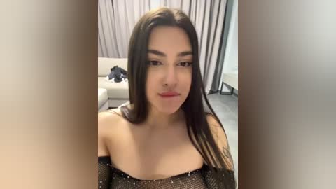 Video of a young woman with long, straight black hair, wearing a glittery black off-shoulder dress, standing in a modern, minimalist living room with light gray curtains and a white couch in the background.