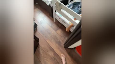Video of a messy bedroom with wooden floor, a white crib, and a bare foot on the floor.