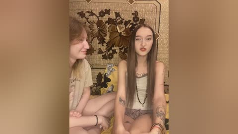 Video of two young women sitting on a bed with floral patterned bedding. One with long brown hair, light skin, and wearing a white tank top; the other with long black hair, fair skin, and a black tank top.