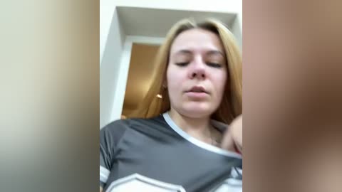 Video of a young Caucasian woman with fair skin and straight, shoulder-length blonde hair, wearing a grey and white striped shirt. She appears calm, with a neutral expression. The background features a beige wall and an open doorway.