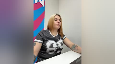 Video of a tattooed, light-skinned woman with shoulder-length blonde hair wearing a grey football jersey, sitting at a desk in a bland office with colorful striped wall art.