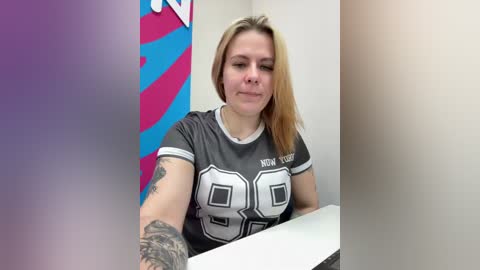 Video of a blonde woman with a tattooed arm wearing a grey \"New York Jets\" jersey, sitting at a white desk in a brightly lit office.