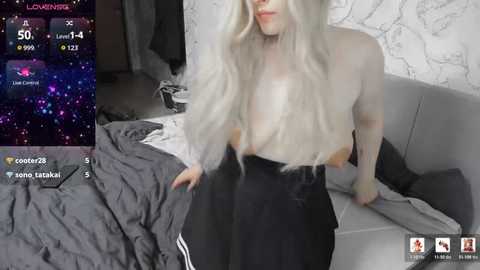 A selfie video of a fair-skinned woman with long, platinum blonde hair, topless, sitting on a grey couch, wearing black pants. The background features a white marble wall and messy bed.