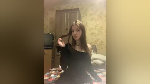 Video of a young woman with long brown hair, wearing a black off-shoulder top, sitting on a bed in a dimly lit, vintage-style room with yellow floral wallpaper and a wooden wardrobe.