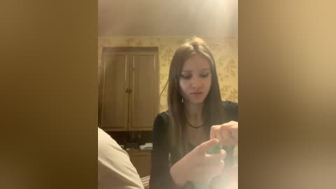 A video shows a young woman with straight, long dark hair and light skin, wearing a black top, holding a green bottle in a dimly lit bedroom with yellow floral wallpaper and wooden cabinets.