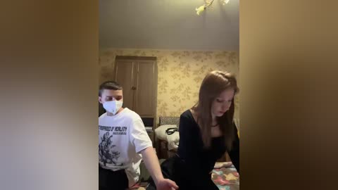 Video of a young woman with long brown hair in a black top, walking towards a man in a white t-shirt, mask, and glasses, in a dimly lit, patterned wallpaper bedroom.
