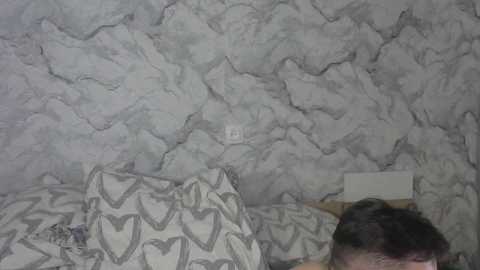 A video of a child's bedroom with a textured gray and white marble-patterned wall. The child, partially visible, lies on a bed covered in a patterned heart pillow and blanket.