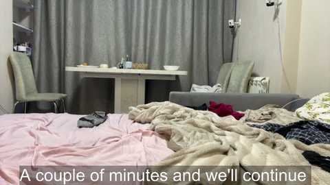 Video of a messy, cluttered bedroom with unmade bed, clothes, and a desk with chairs, grey curtains, and medical equipment in the background. Text overlay: \"A couple of minutes and we'll continue.\