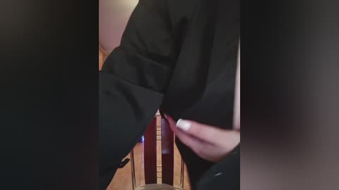 A video of a person in a black robe, possibly a judge, adjusting their robe with their right hand, standing in a dimly lit room with a wooden chair and dark background.