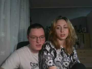 Video of a man with glasses and a white T-shirt and a woman with long blonde hair, both smiling, in a dimly lit bedroom with white curtains and wooden furniture.