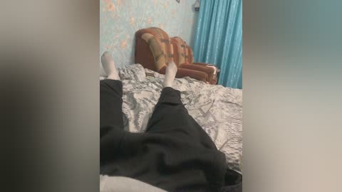 Video of a person lying on a bed with legs crossed, wearing black pants and white socks. The room has blue walls, a patterned headboard, and a turquoise curtain.